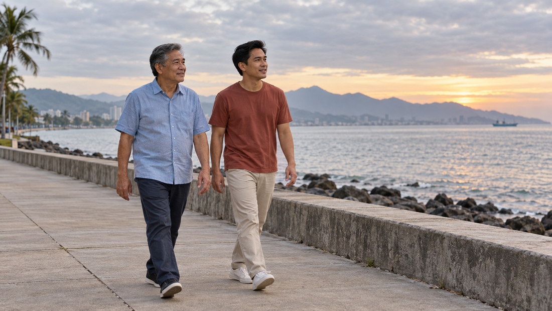 A father and his adult son walk together beside a seawall in soft morning light.