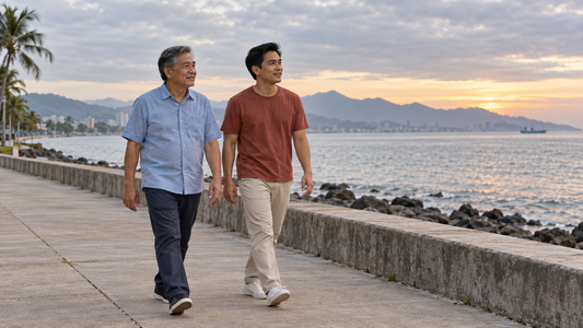 A father and his adult son walk together beside a seawall in soft morning light.